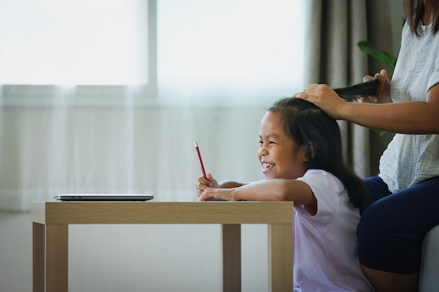 宿題をしているアジアの子供の女の子と母親が居間で髪をとかしている。
