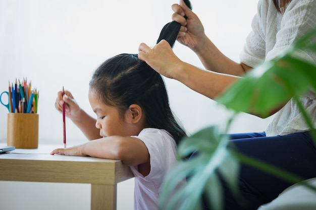 宿題をしているアジアの子供の女の子と母親が居間で髪をとかしている。