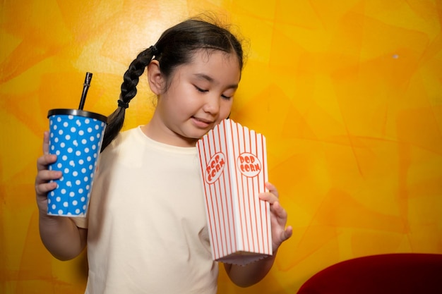 Asian child girl eating popcorn and drinking soda on yellow wall background Cinema concept