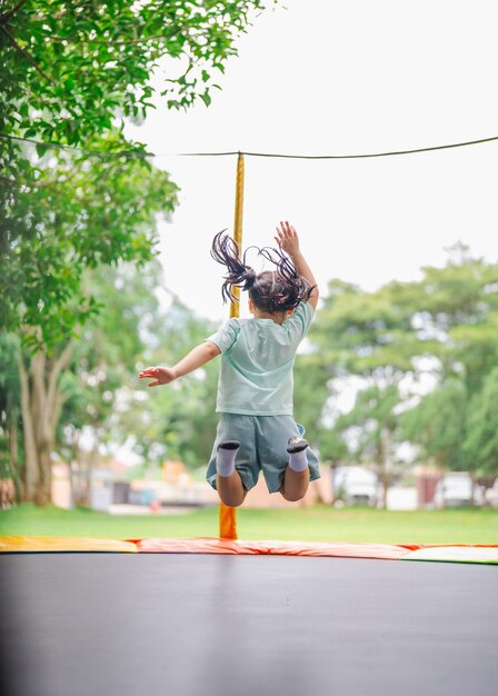 Photo asian child girl is jumping on trampoline on playground background happy laughing kid outdoors in the yard on summer vacation jump high on trampoline activity children in the kindergarten school