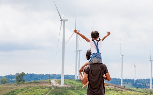 asian child girl riding on father's shoulders in the wind turbine field