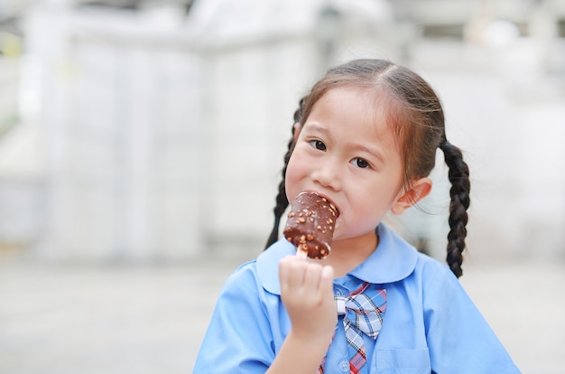 La ragazza asiatica del bambino in uniforme scolastica gode di di mangiare il gelato alla vaniglia saporito del cioccolato.