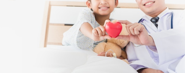 Asian child's hand and male doctor hold red heart lying on bed in the hospital.