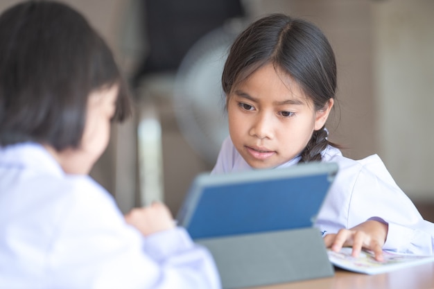 アジアの子供たちの学生の友人が学校に戻って、デジタルタブレットを使用して一緒に勉強し、Covid-19ロックダウン後に学校でノートに書き込みます