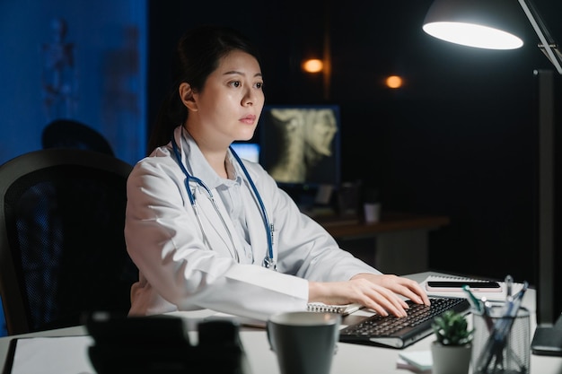 病院のオフィスでコンピューターを使って机の上で働くアジアの中国人女性医師。医療医療患者データ レコード分析の概念。若い女の子の診療所のスタッフが夜に問題を入力して返信する