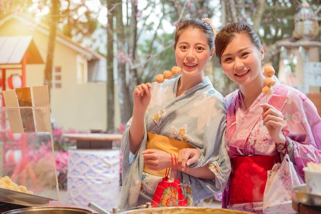 asian chinese girlfriends travel in japan eating fried meatballs skewer together on the local food store and dressed in kimono in the afternoon with sunshine in vintage retro film color.