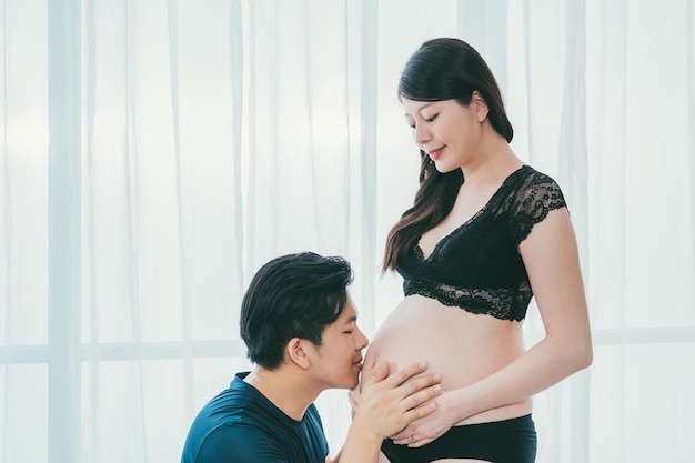 Asian chinese man smiling kissing pregnant wife belly with\
love. young future parenthood couple. male dad holding abdomen of\
sexy mom in black lingerie and pantie by window on happy fathers\
day.