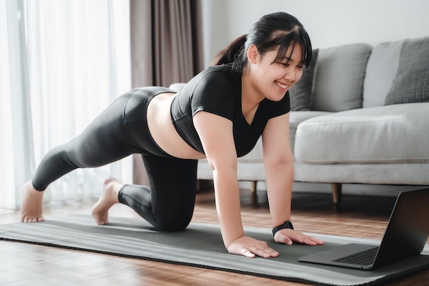 Donna paffuta asiatica seduta sul pavimento in soggiorno pratica lezione di yoga online con il computer