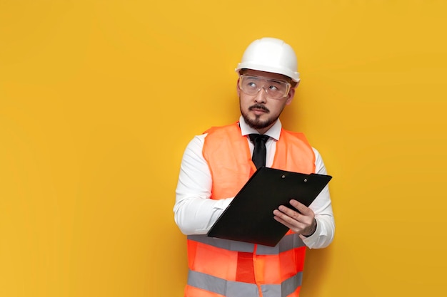 Asian civil engineer in uniform writes plan on paper and looks away on yellow isolated background