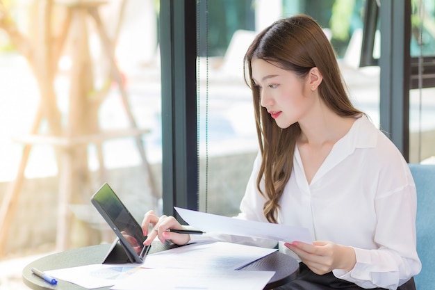 白いシャツを着たアジア人の自信のある女性はタブレットを使って仕事をしていて、テーブルにはたくさんの書類があります。自宅の作業室で笑顔で彼女の顔。