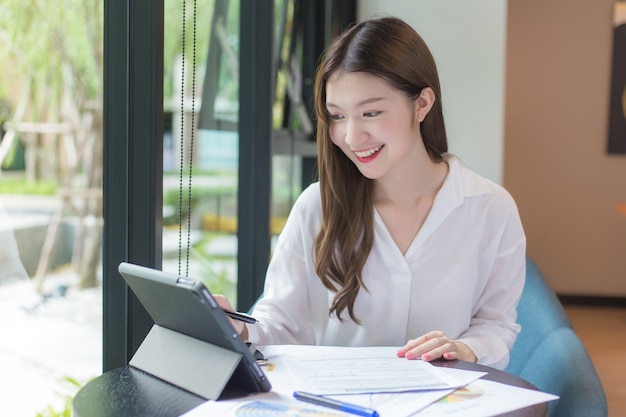 Asian confident woman wear a white shirt is using a tablet to work and there are many documents