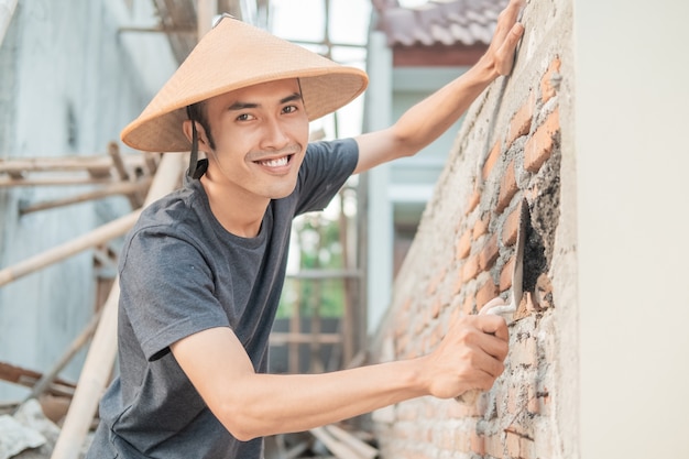 アジアの建設労働者は、スクープを使用してレンガにセメントを広げながら、帽子をかぶってカメラに微笑む