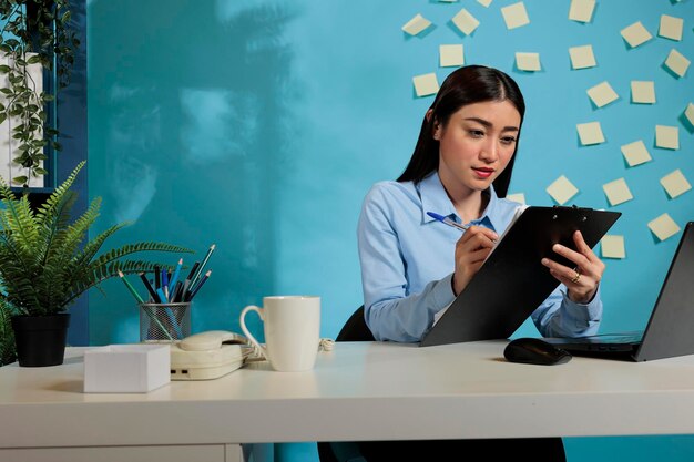 현대 사무실 책상 위에 노트북을 놓고 사무실에서 메모를 하는 아시아 기업 직원. 전문 여성은 완료할 작업 목록을 만드는 프로젝트 마감일을 맞추는 데 집중했습니다.
