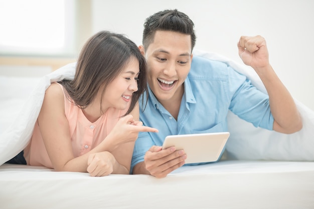 Asian couple lovers enjoy cheer up online esport on smart tablet on holiday vacation in bedroom.