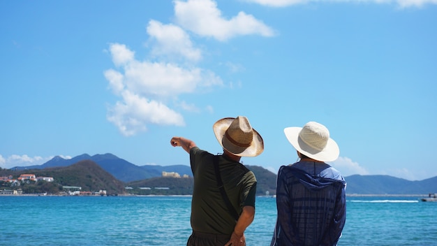 Asian Couple Man and Woman in hats staying on Beach seaside and look into the distance. A man indicates the direction of the hand.