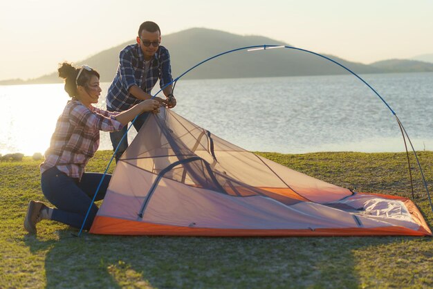 アジアの夫婦が夕暮れの後ろに湖がある芝生でキャンプするテントを準備している