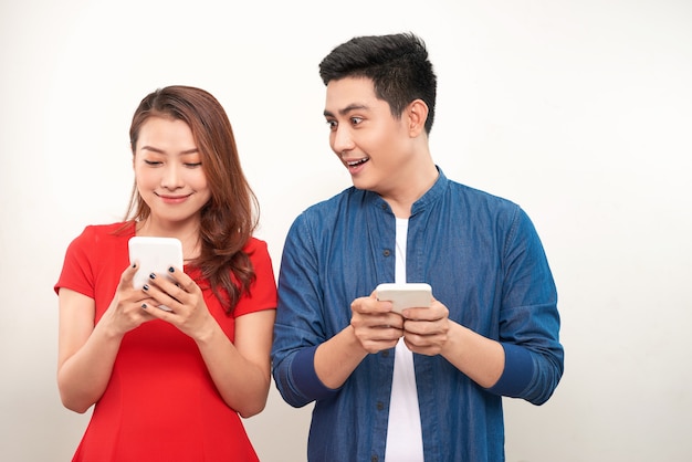ガジェットを使用しているアジアのカップル：彼女のボーイフレンドが彼女の隣に立って、彼女のスマートフォンの画面を見下ろしている間に携帯電話でメッセージを入力しているかわいい女の子