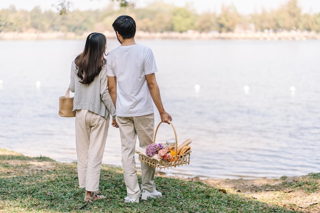 アジアのカップルがピクニック バスケットを持って庭を歩いて恋をしているカップルが屋外の公園でピクニックの時間を楽しんでいます。