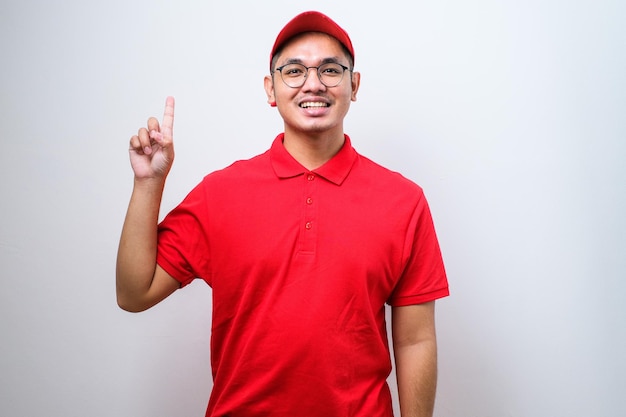 Corriere asiatico che indossa l'uniforme di consegna e il cappuccio che mostra e punta verso l'alto con le dita numero uno mentre sorride fiducioso e felice