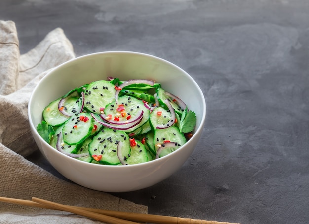 Asian cucumber salad with red onion, chilli pepper and black sesame in white bowl on gray concrete.