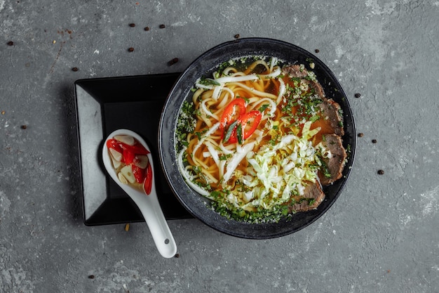 Фото Азиатская кухня, говяжий суп fo в черной тарелке на темноте. суп fo с острым говяжьим бульоном, говяжьей вырезкой, лапшой удон, пастой чили, пекинской капустой, перцем чили, кинзой, соусом ассорти