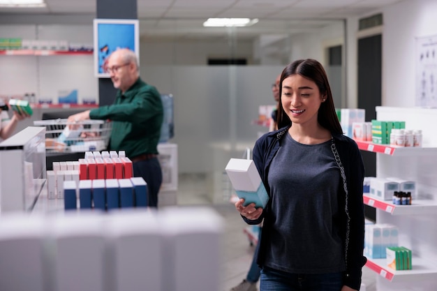 Asian customer holding pills package reading presription before\
buy it. woman walking between shelves shopping for supplements,\
vitamin, medication in pharmaceutical shop. health care\
products