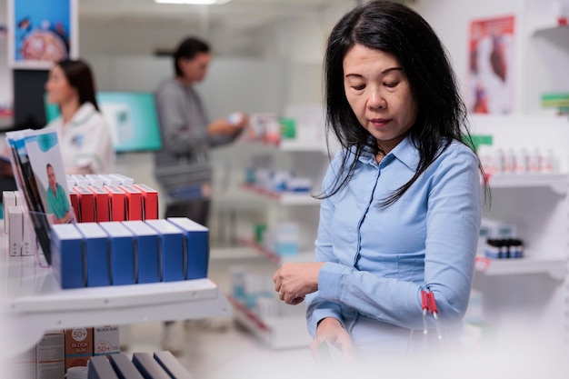 Foto cliente asiatico che prende scatole di integratori dagli scaffali delle farmacie, acquista prodotti farmaceutici e cura delle malattie. donna che guarda pacchetti di medicinali per acquistare forniture sanitarie.