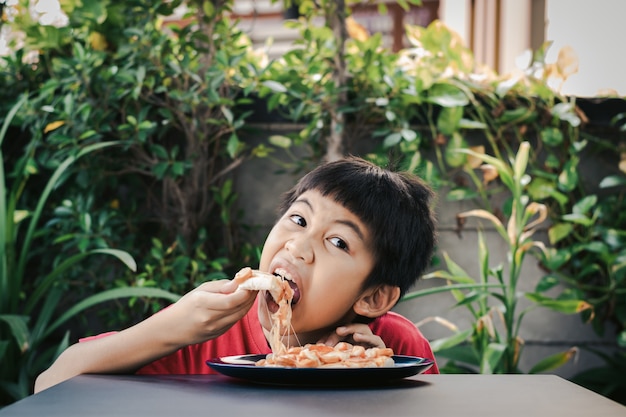 Ragazzo asiatico carino in camicia rossa felicemente seduto a mangiare pizza con sfondo di piante