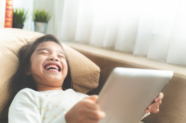 アジアのかわいい子供がタブレットで遊んでいて、家でソファに座っている間に笑う