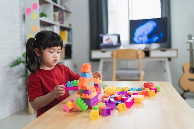 Asian cute funny preschooler little girl in a colorful shirt\
playing with lego or construction toy blocks building a tower in\
kindergarten room or living room kids playing children at day\
care