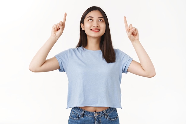 Asian cute girl contemplating interesting scenery pointing index fingers up raising head intrigued smiling broadly observe cool promo advertisement standing white background curious