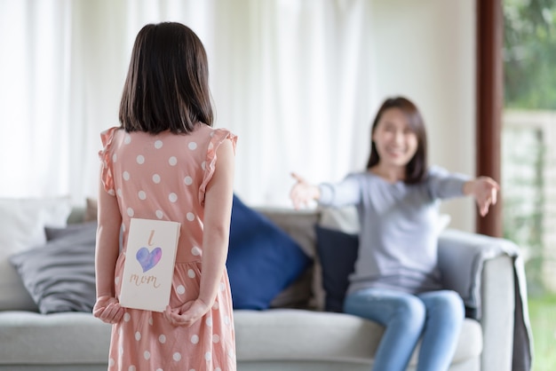 아시아 귀여운 소녀는 집에서 그녀의 어머니를 놀라게 하기 위해 엄마 단어를 좋아하는 손으로 만든 인사말 카드를 숨깁니다.