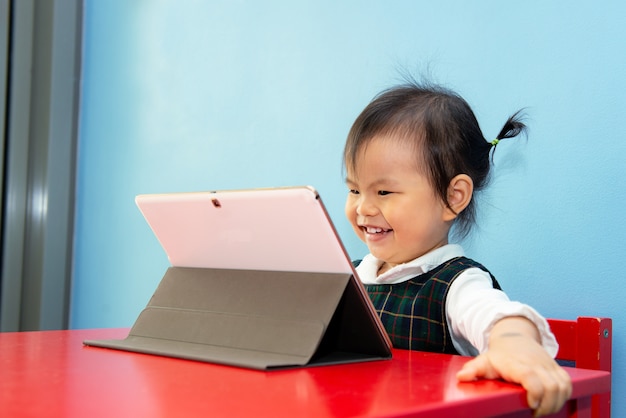 Photo asian cute girl sitting on red chair looks at white tablet, cute baby girl enjoy tablet on