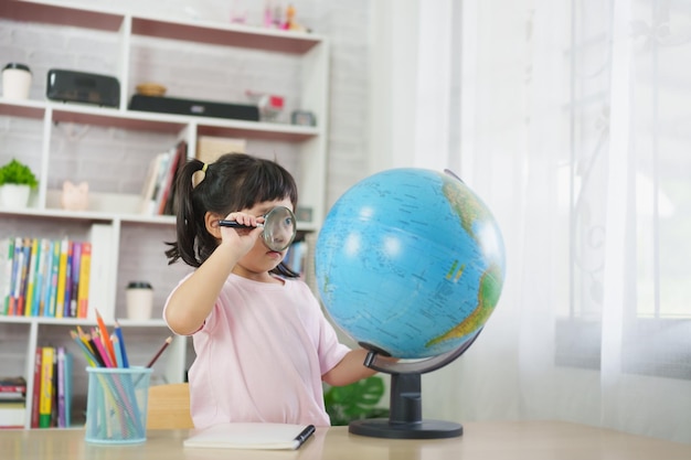 가정 학습 교육 개념의 나무 탁자 내부에 돋보기를 사용하여 교육 지구 모델을 관찰하고 가리키며 웃고 있는 아시아의 귀여운 소녀
