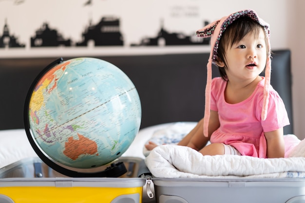 Asian cute little baby girl wearing hat sitting on travel bag with smile feeling funny and laughing on bed in bedroom with world globe put on other side of suitcase bag.