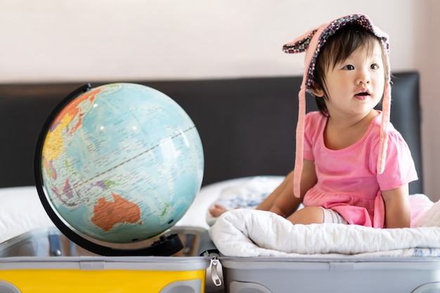 Foto cappello d'uso della piccola neonata sveglia asiatica che si siede sulla borsa di viaggio con il sorriso che ritiene divertente e che ride sul letto in camera da letto.