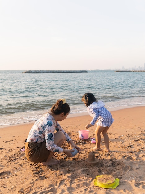 Asian cute little girl and her mother playing or making sand\
castle or digging with sand on tropical beach children with\
beautiful sea sand blue sky happy kids on vacations seaside on the\
beach