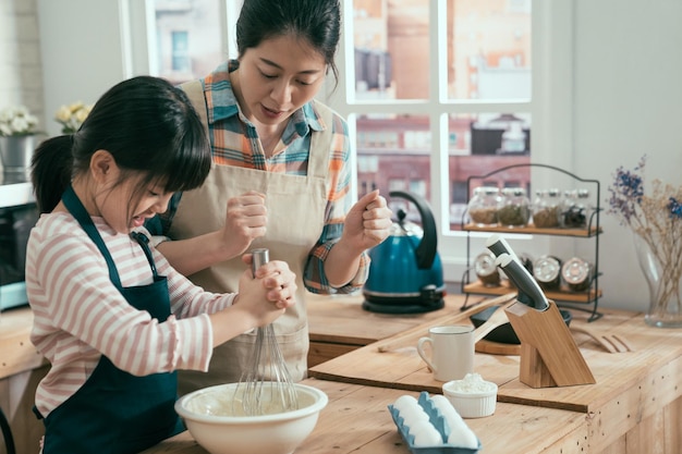 幸せなママが彼女の元気を教えて笑顔で見ながら卵を鞭打ちするアジアのかわいい女の子。娘は幸せな母の日に手作りのケーキを望んでいます。親と子のDIYはキッチンで時間を焼きます。