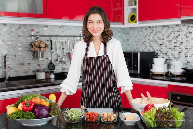 Asiatica carina donna di mezza età in un grembiule in piedi sulla moderna cucina nuova tonalità di rosso con varie verdure e ingredienti per la preparazione di insalate. concetto per casalinga e madre di stile di vita.