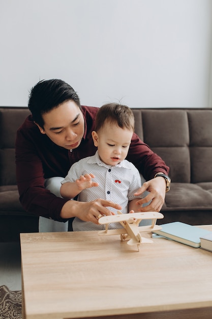 Asian dad and his cute little son playing with a wooden plane in a bright room