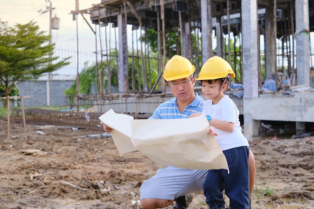 Photo asian dad and son wearing yellow construction helmet standing in front of construction site