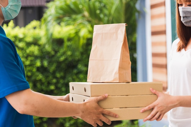 Asian delivery express courier young man giving paper bags fast food and pizza box to woman customer receiving both protective face mask, under curfew quarantine pandemic coronavirus COVID-19