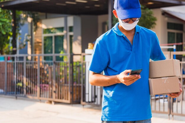 Asian delivery man courier online holding deliveries out boxes and using mobile phone contact the customer he protective face mask service under curfew quarantine pandemic coronavirus COVID-19