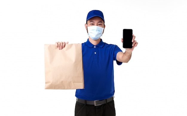 스마트 폰 흰색 배경에 고립 된 종이 가방을 들고 파란색 유니폼에 얼굴 마스크를 착용하는 아시아 배달 남자. covid19 동안 빠른 배달 서비스.