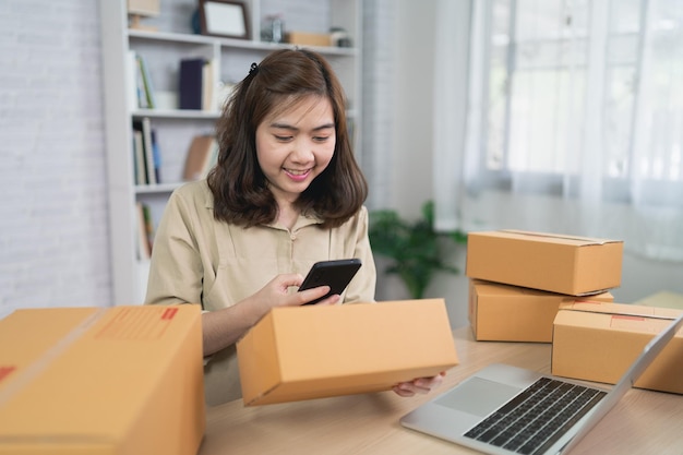asian delivery woman freelance worker smiling using mobile scan barcode for boxes for sending or conveying parcels by mail work at home