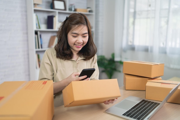 Lavoratore freelance della donna asiatica delle consegne che sorride utilizzando il codice a barre di scansione mobile per le scatole per l'invio o il trasporto di pacchi per posta lavoro a casa