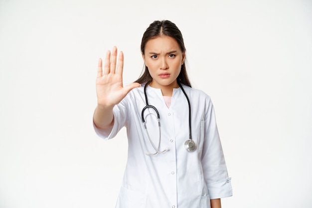 Азиатский врач показывает жестом остановки ладони, серьезная недовольная женщина-врач протягивает руку, запрещает действовать ...