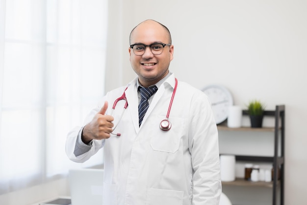 Asian doctor standing posing showing thumbs up work from home Positive male physician with stethoscope looking at camera in hospital background Healthcare and medical concept