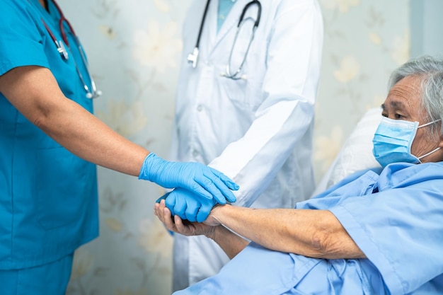 Asian doctor team and old senior lady patient stack hands together as team for motivation new normal to treat infection Covid19 Coronavirus at hospital ward