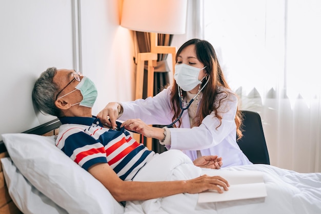 Asian doctor using a stethoscope to examines a senior man at home The doctor checking up and consulting for retirement health partienthospital services and wearing a mask to protect covid19